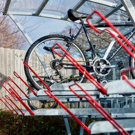 A Two Tier Bike Rack holds several bicycles in an outdoor area. The Chelsea Cycle Shelter is made of metal with a transparent roof. One black bike is parked on the upper level, and a blue and black bike is on the lower level. Trees and a wooden fence are in the background.