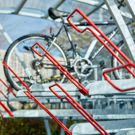 A close up of a Two Tier Bike Rack holds several bicycles in an outdoor area. The Chelsea Cycle Shelter is made of metal with a transparent roof. One black bike is parked on the upper level, and a blue and black bike is on the lower level.