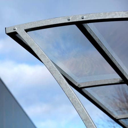 A close-up photo of the Chelsea Cycle Shelter a modern metal and glass canopy structure, viewed from below against a backdrop of a cloudy sky and blurred building. The structure features a brushed metal frame and transparent glass panels.