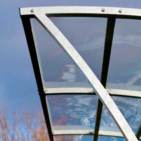 A close-up photo of the Chelsea Cycle Shelter a modern metal and glass canopy structure, viewed from below against a backdrop of a cloudy sky and blurred building. The structure features a brushed metal frame and transparent glass panels.