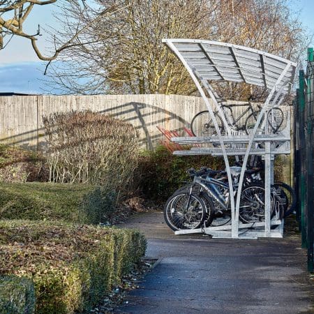 A Two Tier Cycle Rack within a covered Chelsea Shelter in an outdoor area next to a metal fence. Several bicycles are parked in the rack, with a few on the upper tier of a two-level storage system. The ground is paved, and there are bushes and buildings in the background.
