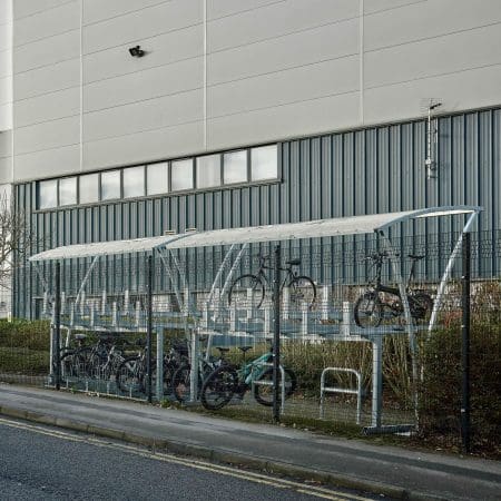 A Two Tier metal bike rack stands against a large industrial building. The roofed structure shelters several parked bicycles on both the upper and lower levels. The area is enclosed with a metal fence and a small hedge runs along one side.
