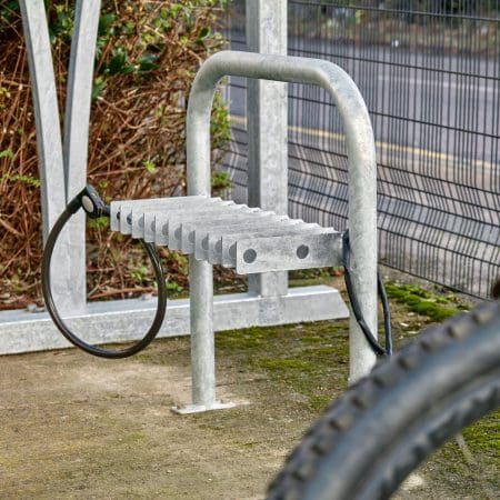 A metal bike rack with a lockable loop and grooved slots is installed on a concrete surface. A bicycle tire is partially visible in the foreground. The background shows wire fencing, shrubbery, and a street.