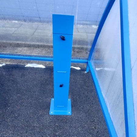 Close up of a light blue e-bike charging stand installed outside in a polycarbonate e-bike charging shelter with snow in the background on the ground