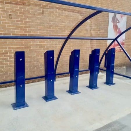 A row of blue e-bike charging stands installed outside under a blue polycarbonate bike shelter, in front of a brick wall