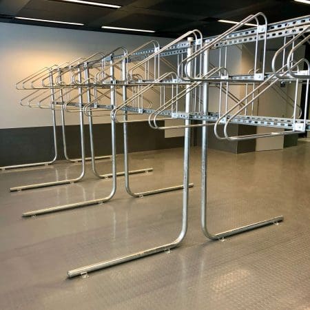 A row of empty, metal bike racks in a modern, indoor setting with grey flooring and white walls. The racks are lined up neatly in parallel, with multiple hooks but no coats or other items hanging from them. The room appears clean and well-lit.