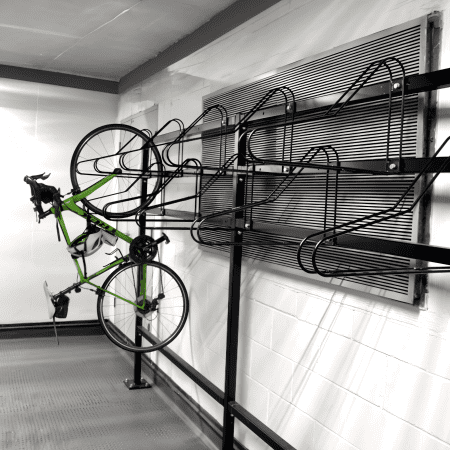 A green road bike is hanging vertically by its front wheel on an industrial-style bike rack attached to a white brick wall. The bike rack is made of black metal and includes several curved hooks for multiple bicycles. The flooring is gray and the setting appears to be a garage or storage area.
