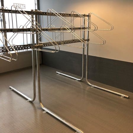 A metal bike rack stands in an empty room with several wire hangers evenly spaced on its rods. The room has a grey tiled floor and a partially visible window, allowing some natural light to enter. The walls are painted in two tones, grey on the bottom and white on the top.
