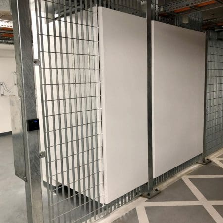 A secure bike storage area with metal wire fencing and a closed gate. The walls are white, and the floor is concrete.