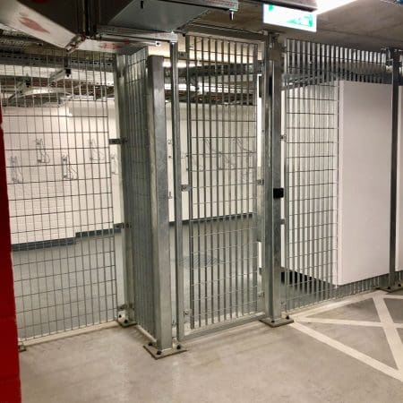 A secure storage area with metal wire fencing and a closed gate. The walls are white, and the floor is concrete. There are metal support beams and pipes visible overhead. The area appears to be part of a larger facility, possibly a warehouse or storage room.