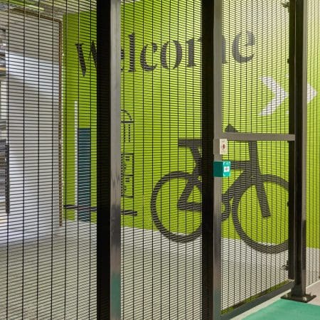 Our high security mesh fencing protecting a storage area with a green wall displaying the word "welcome" in large letters. The wall also features a graphic of a bicycle and directional arrows. The area appears to be secured with metal bars and has a locked gate.