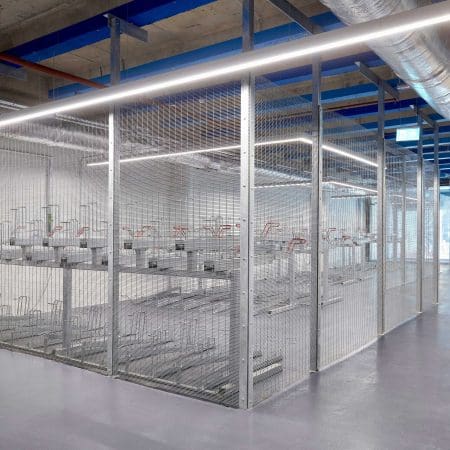 A large, well-lit room with industrial decor containing several rows of metal two tier bike racks enclosed by the anti-climb mesh fencing. The ceiling features exposed pipes and beams, and the floor is a smooth, polished concrete surface.