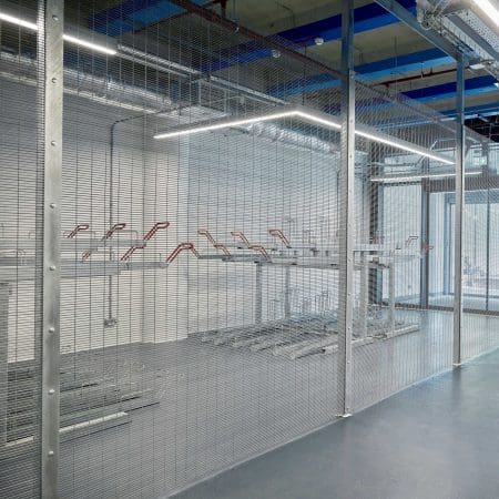 The image shows a secured indoor bicycle storage area with anti-clim mesh fencing. Inside, empty bike racks are arranged along the walls under overhead fluorescent lighting. The flooring is smooth and grey, and there are visible metal pipes and structural beams on the ceiling.