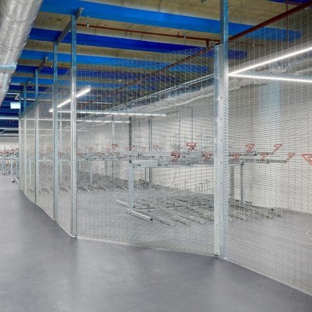 A secured indoor bike storage area with metal mesh fencing and two-tier bike racks. The space has a concrete floor, and the ceiling features blue beams and industrial piping. Fluorescent lights provide bright illumination.