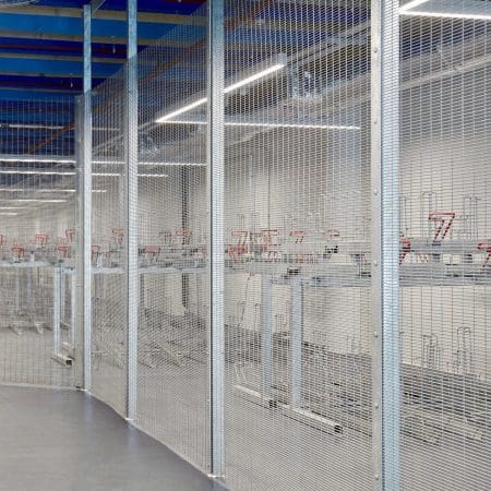 A spacious, well-lit industrial storage area with several metal bike racks mounted on the wall behind a mesh wire fence. The ceiling has noticeable blue beams and the floor is a polished, grey concrete.