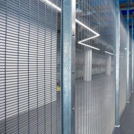A secure indoor area with a security mesh fence and sturdy steel beams, featuring an empty corridor with polished concrete floors and a ceiling with linear LED lighting. The atmosphere appears industrial and modern.