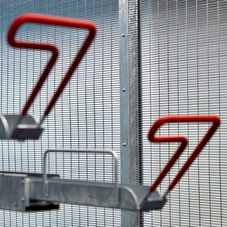 Close-up of a metallic bike rack with curved red hooks against a background of wire mesh fencing. The bike rack's design features sleek, angled red components that stand out against the industrial aesthetic of the grey mesh.