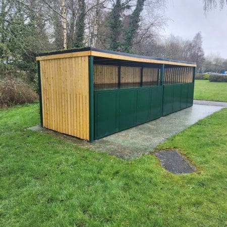 A long outdoor wooden structure with a black roof and green lower panels, set on a concrete slab surrounded by grass. Designed for secure bike storage, it features weather-resistant materials and is located in a park-like area with trees and a cloudy sky in the background.