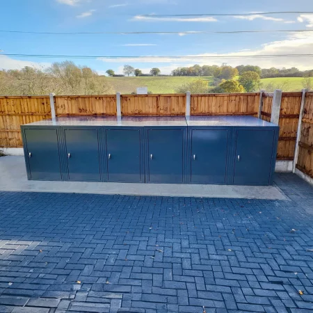 A row of weather-resistant, dark grey metal bike storage lockers is positioned outdoors on a paved surface in front of a wooden fence. These secure bike lockers provide peace of mind while you enjoy the scenic view of green fields and a partly cloudy sky in the background.