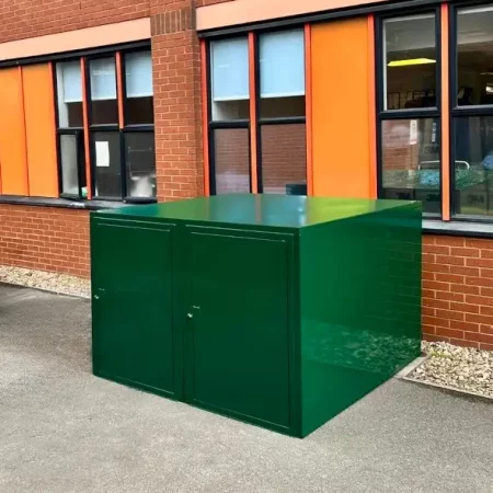 A large green metal storage box, functioning as a secure bike storage unit, is situated outside a brick building with several windows featuring orange panels. This weather-resistant bike store is placed on a paved area next to a round concrete planter filled with greenery.