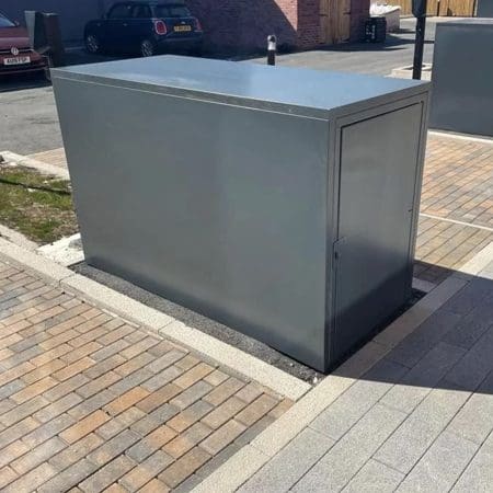 A rectangular gray metal box, featuring a three-point locking system and designed for secure bike storage, is positioned on a paved area that includes light gray and brown bricks. Several identical weather-resistant boxes are also visible in the background. The surrounding area shows a partial view of a parked car, a brick wall, and a wooden fence.
