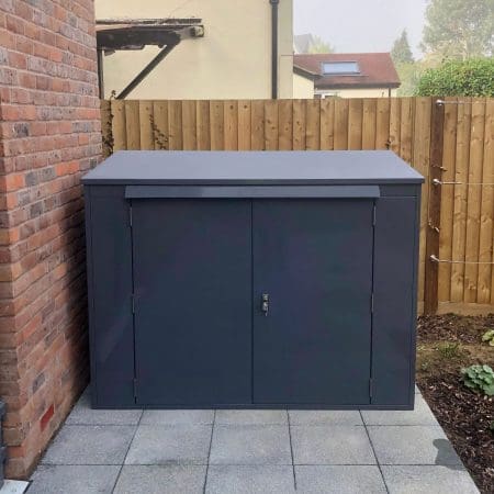 A horizontal bike locker that is positioned on a tiled area adjacent to a brick wall and wooden fence. The shed has a flat top and two front doors with a lock. It is placed in an outdoor area with some green shrubs visible in the background.