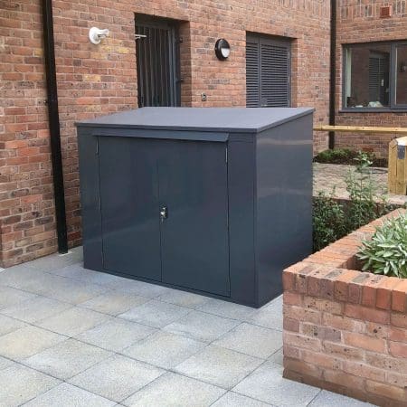 A sleek, dark-colored horizontal bike locker with a sloped roof stands on a tiled patio next to a brick wall of a modern building. The shed has double doors with a locking mechanism and is positioned near a small garden area with green plants.