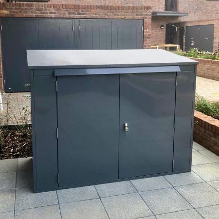 A gray metal horizontal bike locker with two front doors and a lock is placed on a tiled patio area. The box is situated against a brick building, with windows visible in the background.