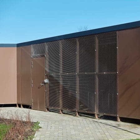 A large brown Carlton Shelter bike enclosure stands prominently beside a brick walkway, inviting visitors to explore its surroundings.