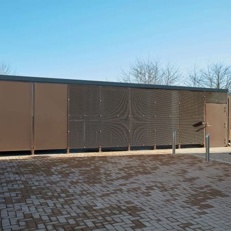 A large brown Carlton Shelter bike enclosure stands prominently beside a brick walkway, inviting visitors to explore its surroundings.