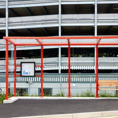 Bus stop with a red metal structure and a transparent roof, situated in front of a multi-level parking garage. An information board is mounted within the shelter. The pavement is freshly paved, and some greenery can be seen at the edges.