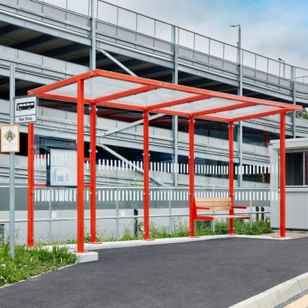 A modern bus stop with a bright red frame and a glass roof stands next to a fenced parking structure. The area is clean with a smooth asphalted surface, and a wooden bench is situated under the shelter. A bus stop sign is visible on the left.