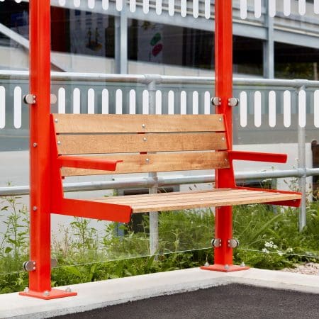 A wooden bench with red metal supports is placed in a modern outdoor setting, featuring a glass barrier and a building with a white fence in the background. The bench has armrests and is positioned on a neatly paved area surrounded by some greenery.