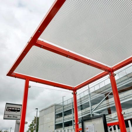 A bus stop with a red metal frame and a perforated metal roof. There is a bus stop sign on the left, and a modern building with glass windows in the background. The sky is cloudy.