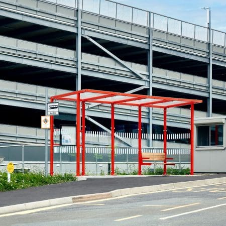 A small, red-framed bus stop with a bench underneath stands in front of a multi-level parking garage. The bus stop is situated on a sidewalk adjacent to a road, with clear blue skies in the background.