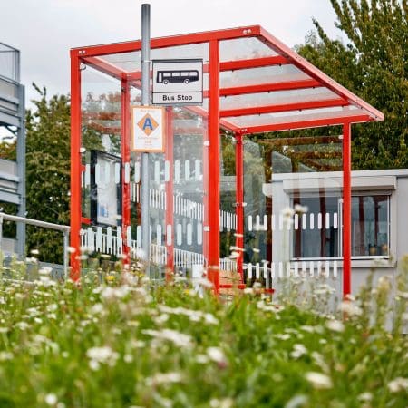 A modern red-framed bus stop with a glass shelter sits in front of a green area with wildflowers. There's a "Bus Stop" sign on top of the shelter and various plants and trees in the background on a cloudy day.