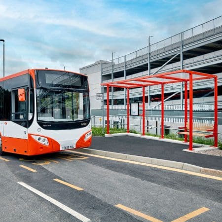 A red and white city bus is parked at a bus stop in front of a multi-story parking garage. The bus stop has a covered shelter with red frame and seating. The scene is under a partly cloudy sky with an empty road beside the bus.