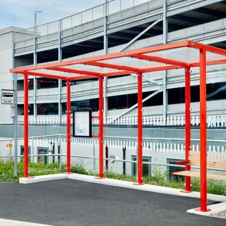 A bright red bus shelter stands on a black pavement before a multi-story parking garage. The shelter, which has a simple modern design and one small bench, is open on four sides and topped with a flat roof. A schedule or map is displayed on one of the side posts.