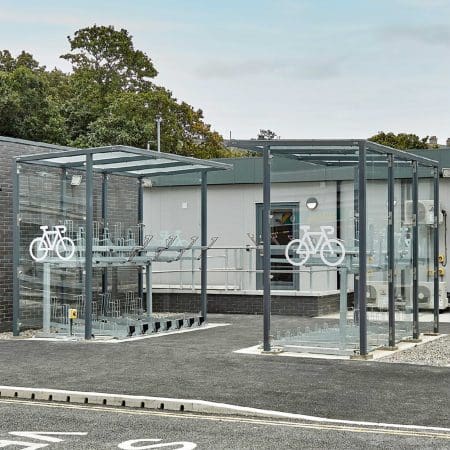 Modern bike parking facility with transparent roofing and sides, equipped with multiple racks and bike symbols, adjacent to a building and parking lot.