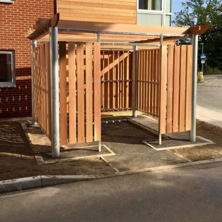 A wood-paneled, open-air pergola bin store with a metal frame, featuring a flat roof and situated on a flat, partially paved area near a red-brick building. The structure appears to be designed for outdoor use, possibly as a storage or seating area.