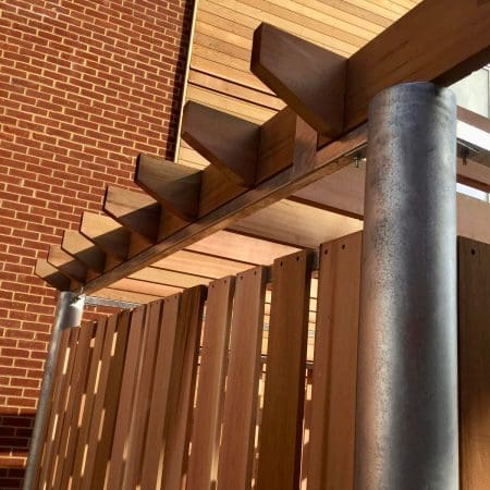 A close-up view of a modern wooden pergola attached to a brick building. The structure features horizontal wooden planks and large metal support posts. Sunlight casts shadows, highlighting the textures of the wood and brick.