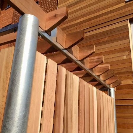 A close-up view of a wooden fence with vertical slats and metallic posts, set against a background of wooden siding and beams. The structure features a blend of natural wood tones and metallic elements, showcasing a modern architectural design.