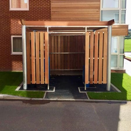 A covered bin store with wooden slats and a metal frame next to a brick and glass building. The shelter has an open front and back, a flat roof, and is surrounded by small patches of artificial grass.