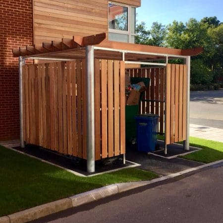 A small outdoor bin store with vertical wooden slats houses green and blue recycling and trash bins. The enclosure has a metal frame and a wooden slatted roof. It is situated between a paved road and a brick building, with a patch of grass on one side.
