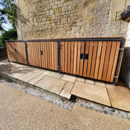 A row of bespoke cycle storage units with metal frames is lined against a stone building. The ground is covered in gravel, and a small cobblestone path is visible on the left side. A tree with sparse foliage is adjacent to the cycle storage units.