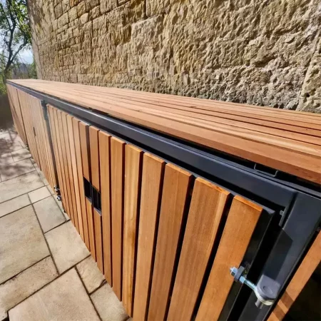 A wooden storage enclosure with a sleek design, perfect for bespoke cycle storage, is set against a stone wall. Sunlight casts shadows on the neatly arranged wooden slats, while the surrounding area features a paved walkway and greenery in the background.