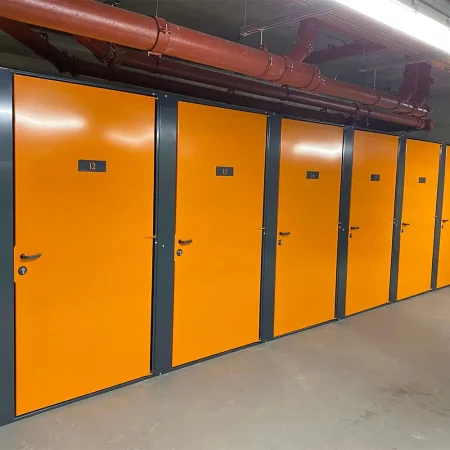 A row of storage units with bright orange doors and dark frames in an industrial setting. Each door has a number label, including 12 and 13 visible. Overhead pipes and fluorescent lighting can be seen in the concrete hallway—ideal for high-security bike storage in residential property development projects.