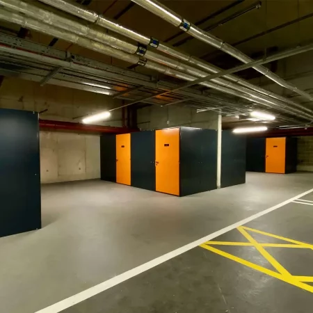 A row of brightly lit storage units in an indoor facility, perfect for storing bikes. Each unit has a door painted in orange with black trim, numbered 10 through 14, and is equipped with a lock. Overhead, there are exposed pipes and a long fluorescent light fixture—ideal for the Tenant Cycle Store's high-security space.