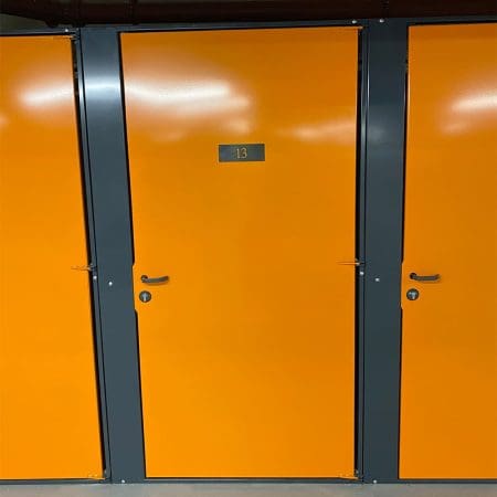 A row of bright orange storage lockers with dark gray frames is perfect for a Tenant Cycle Store, offering a high-security space. The middle locker has a metal sign displaying the number 13. All lockers have black handles and a smooth, shiny finish. The ceiling above has exposed red piping.