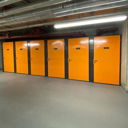 A high-security bike storage area is situated in a corridor with several bright orange doors, numbered, and set in grey frames. The ceiling has exposed pipes and ducts. The floor is clean and smooth, well-lit by overhead fluorescent lights. A directional sign is visible on the left wall.